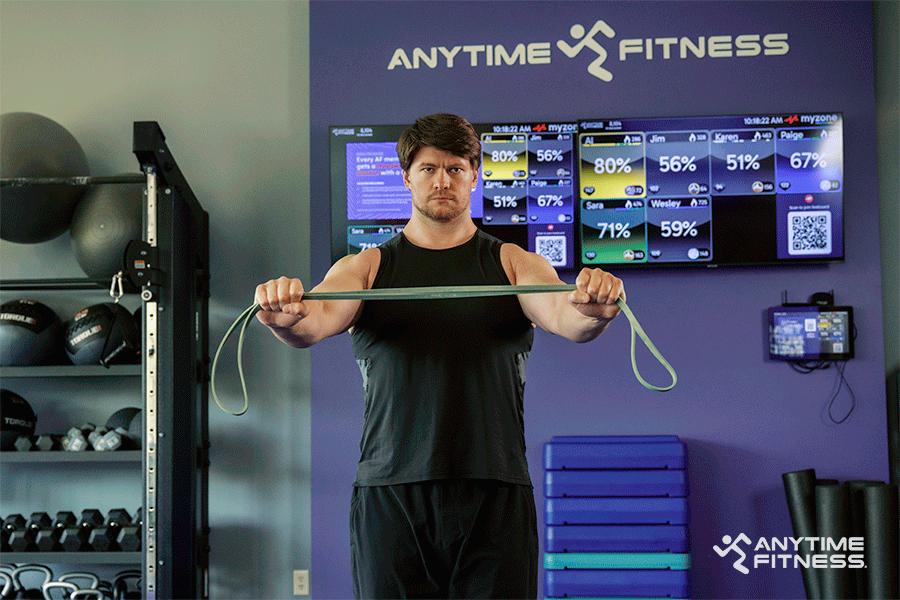 Man in an Anytime Fitness gym demonstrating banded pull-aparts, part of an upper-body warm-up.