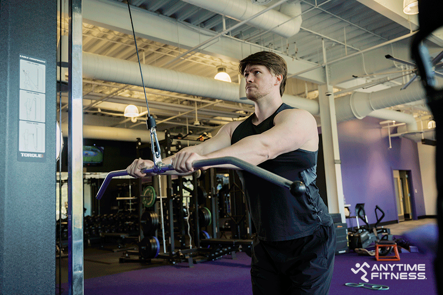 Man in an Anytime Fitness gym performing a straight-arm pulldown during a back and bicep workout.
