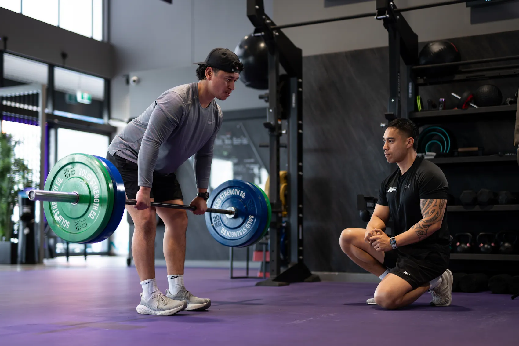 Gym member lifting a loaded barbell while a personal trainer kneels nearby watching his form
