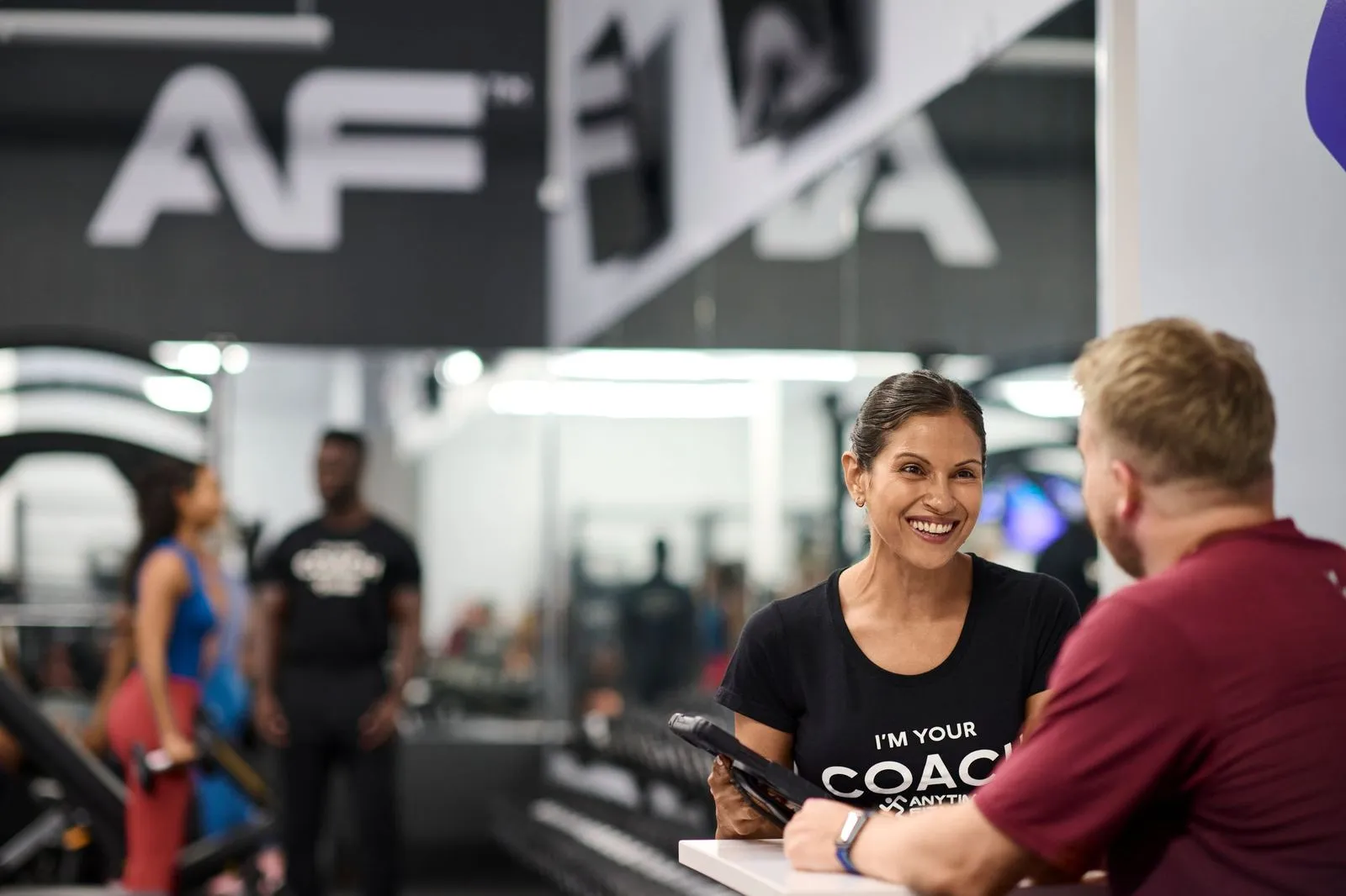 Una entrenadora sonriente con una camisa negra que dice «Soy tu entrenadora» habla con un hombre que lleva una camisa granate en un gimnasio, con personas borrosas haciendo ejercicio de fondo.