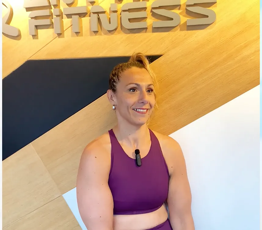 Mujer sonriente con brazos musculosos que lleva un sujetador deportivo morado y un clip para micrófono, sentada frente a una pared de madera con la palabra «FITNESS» en la parte superior de ella.