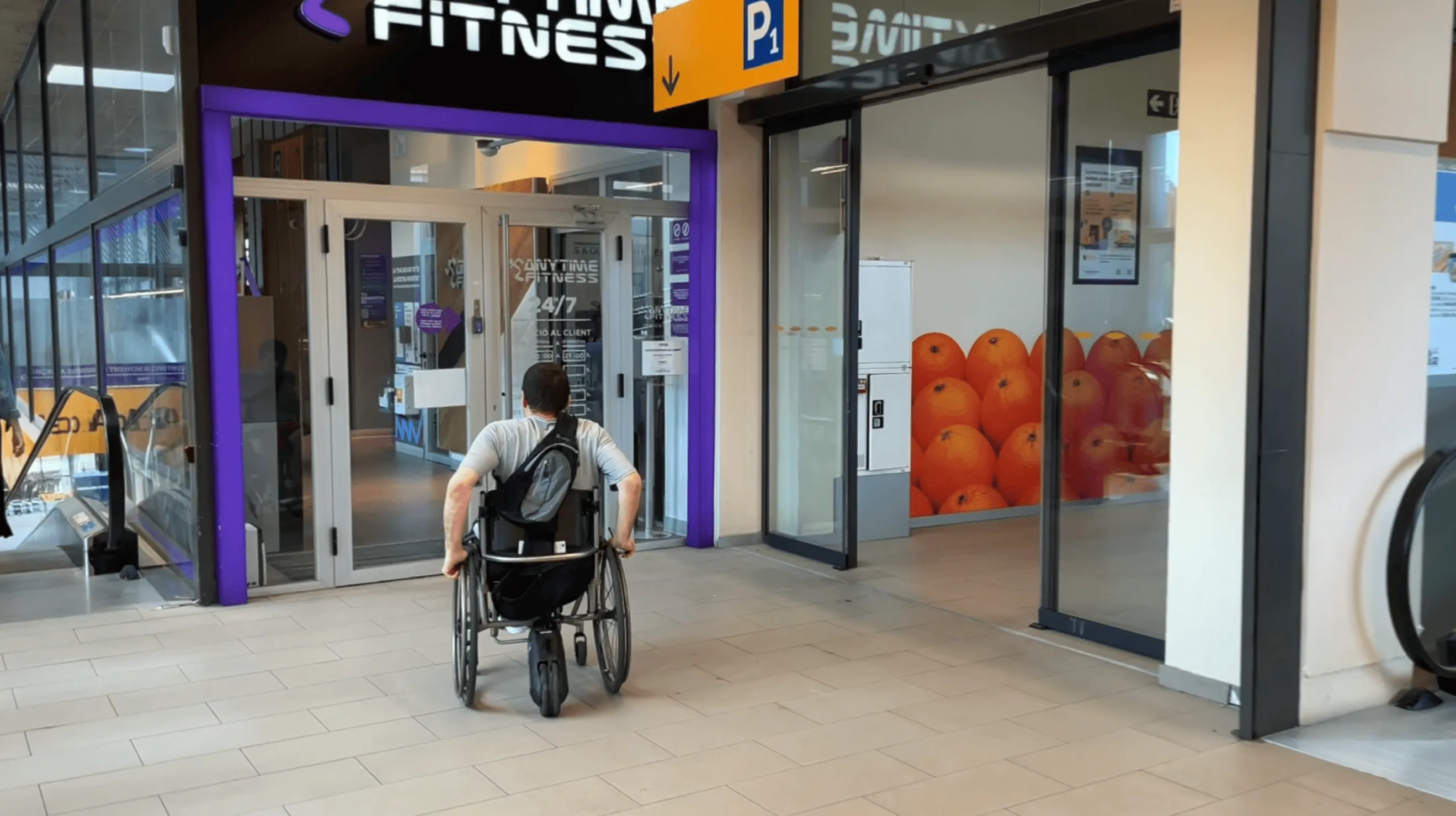 Persona en silla de ruedas que se dirige hacia la entrada de un gimnasio Anytime Fitness con puertas dobles de vidrio y un marco morado en un centro comercial o complejo cubierto.