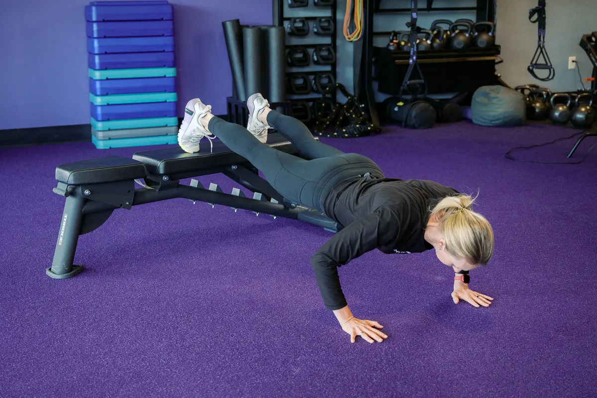 Anytime Fitness Coach demonstrating a feet-elevated push-up using a bench in a gym.