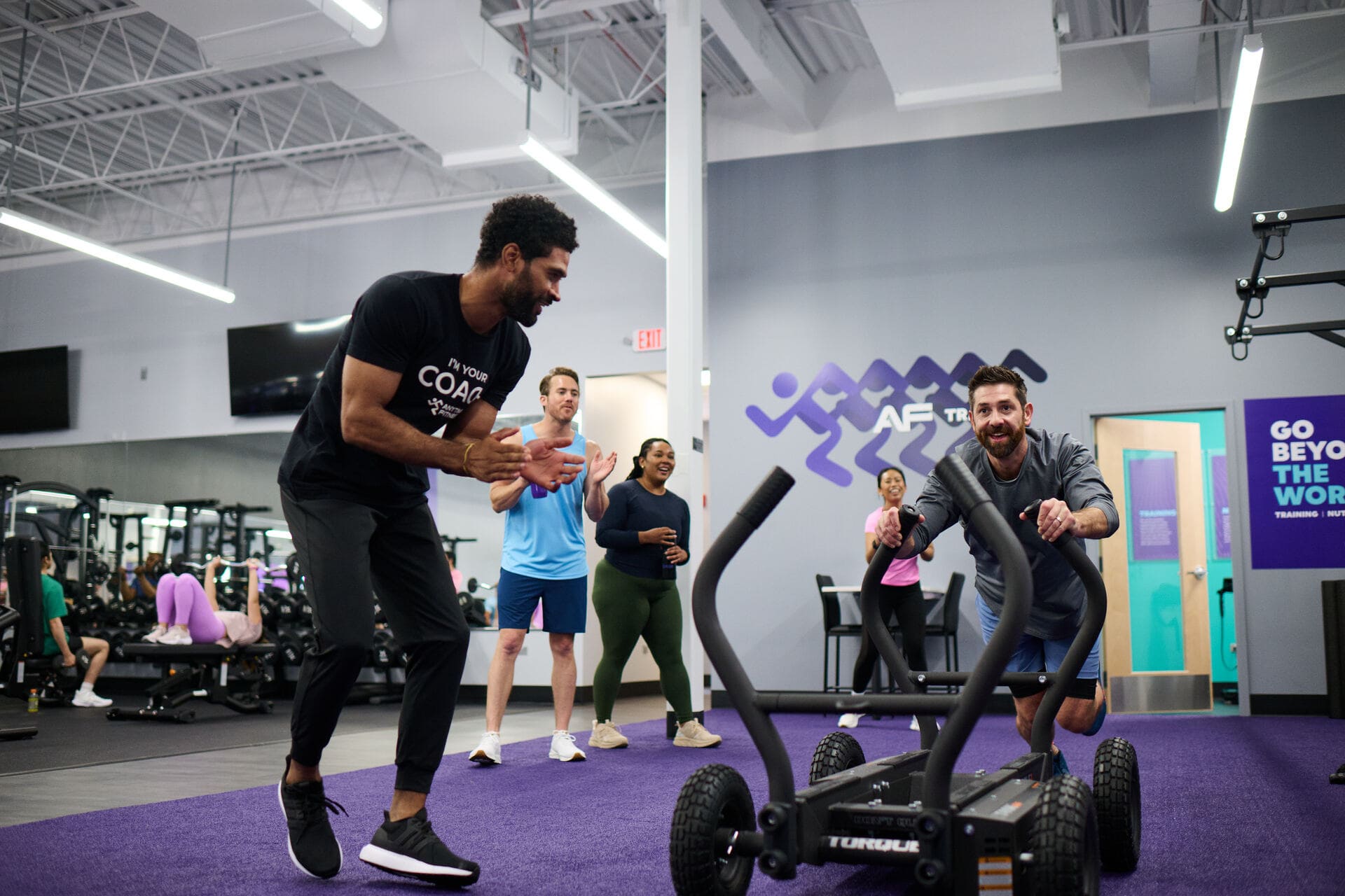 A coach wearing a black shirt that says 'I'm your coach' encourages a man pushing a weighted sled in a gym, while three others watch and cheer them on.