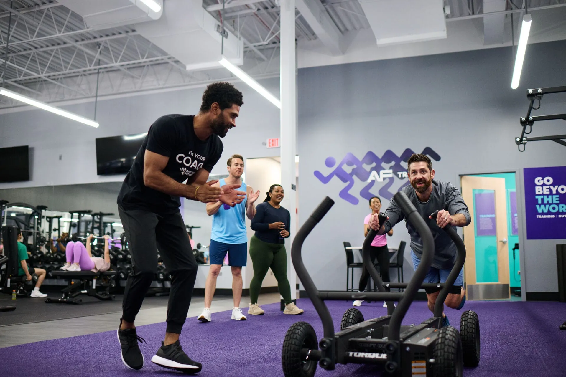 A coach in a black coach shirt encourages a man pushing a weighted sled while three others cheer him on.