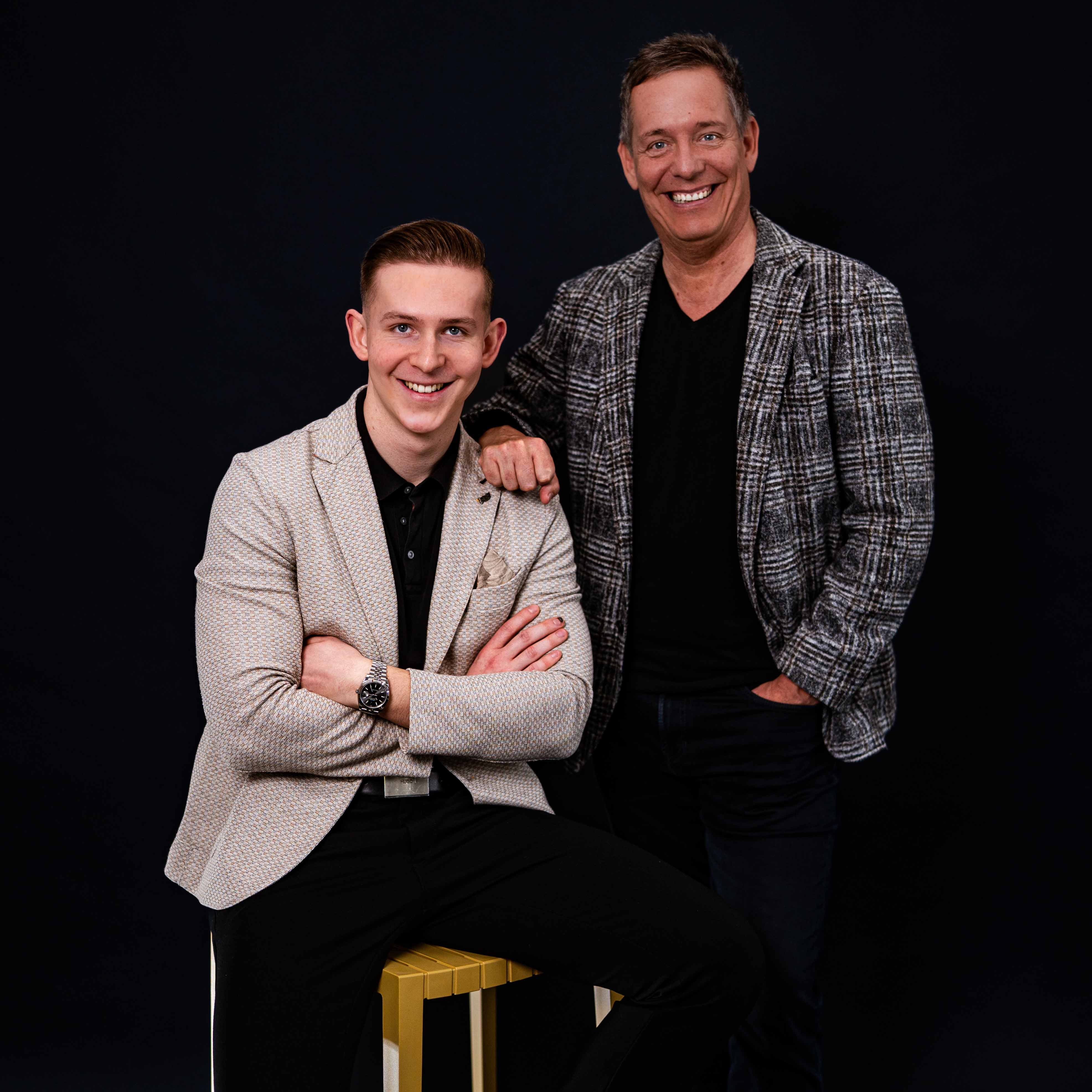 Zwei lächelnde Männer vor schwarzem Hintergrund, einer sitzt auf einem gelben Hocker mit verschränkten Armen, der andere steht daneben mit Hand in der Hosentasche.