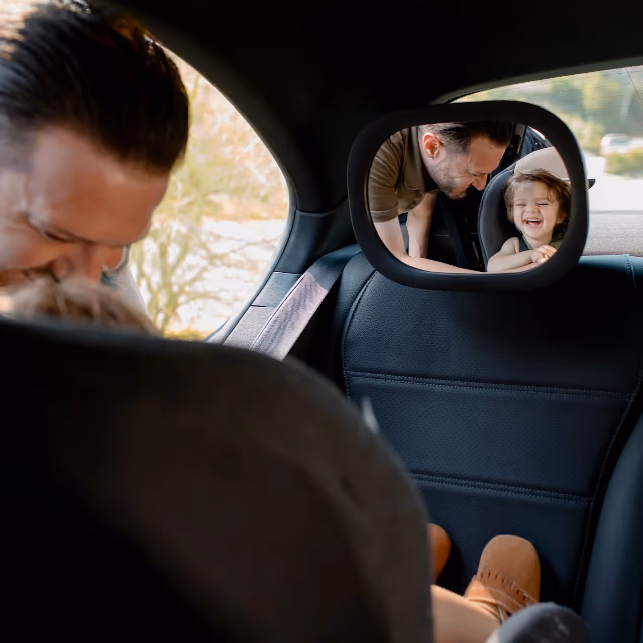 A man laughs with his baby that we can see in the mirror's reflection from the driver's point of view inside a car.