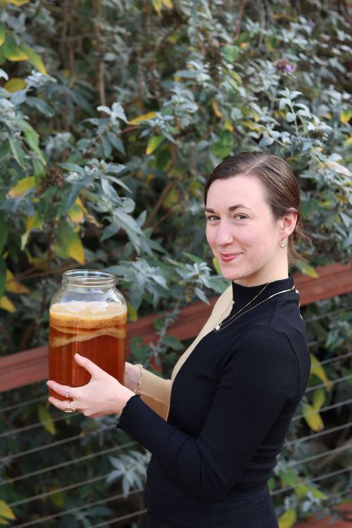 Person holding a large jar of kombucha with a SCOBY, standing outdoors in front of green foliage, during a kombucha brewing session