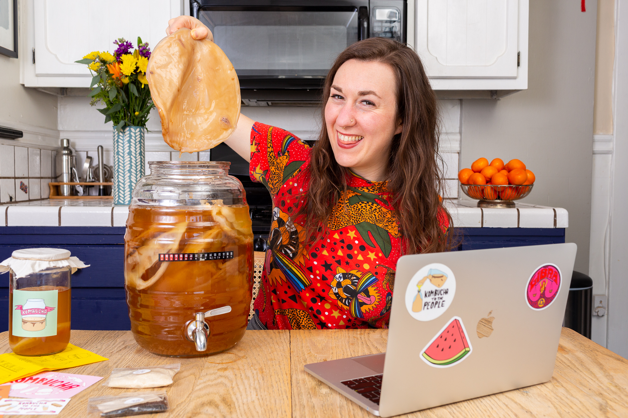 Smiling woman holding a large SCOBY above a glass jar of kombucha on a kitchen table with a laptop and kombucha supplies.