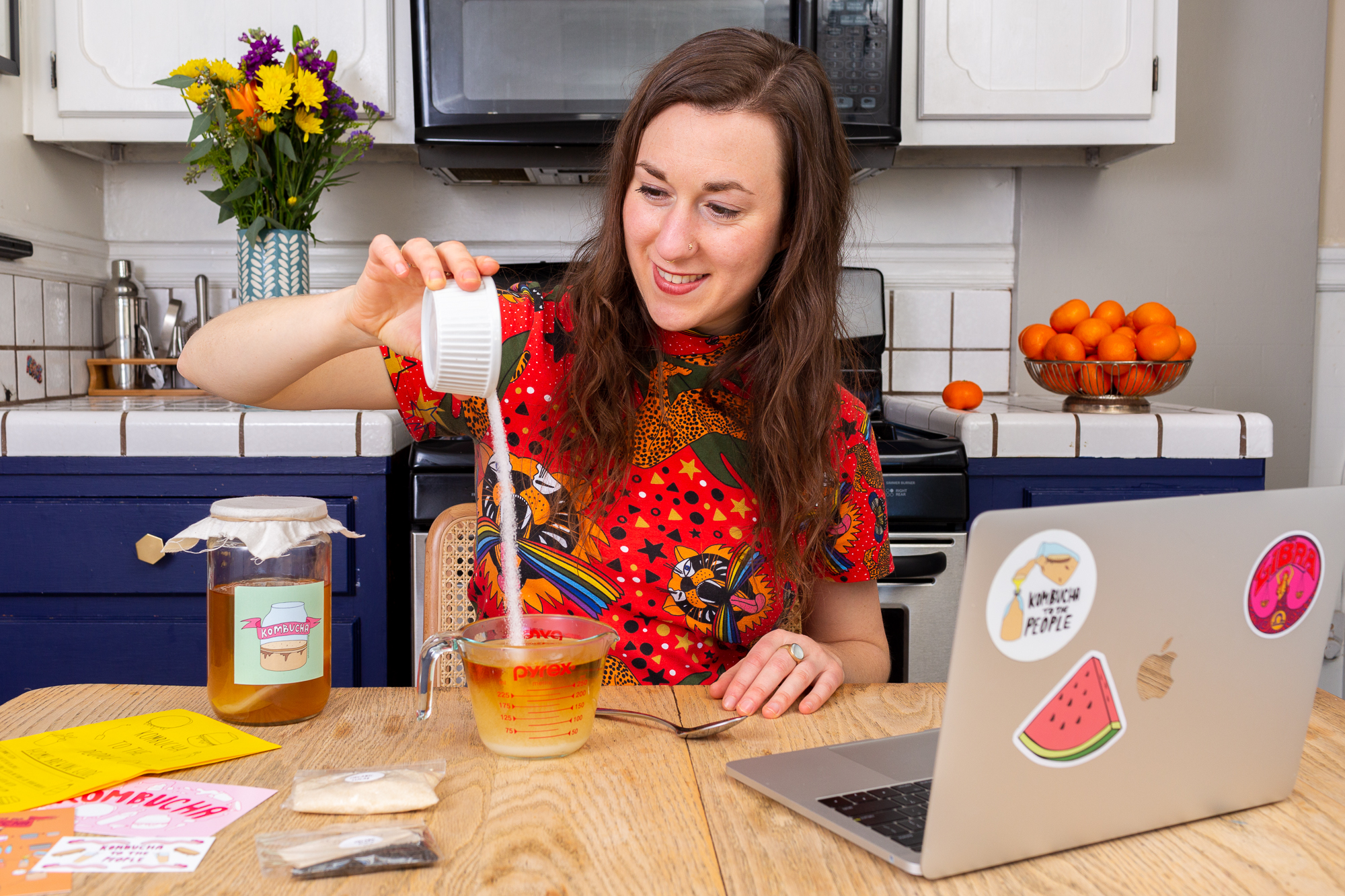 Smiling woman in a colorful shirt pouring sugar into a glass measuring cup on a wooden table with kombucha supplies and a laptop covered with stickers in a kitchen.
