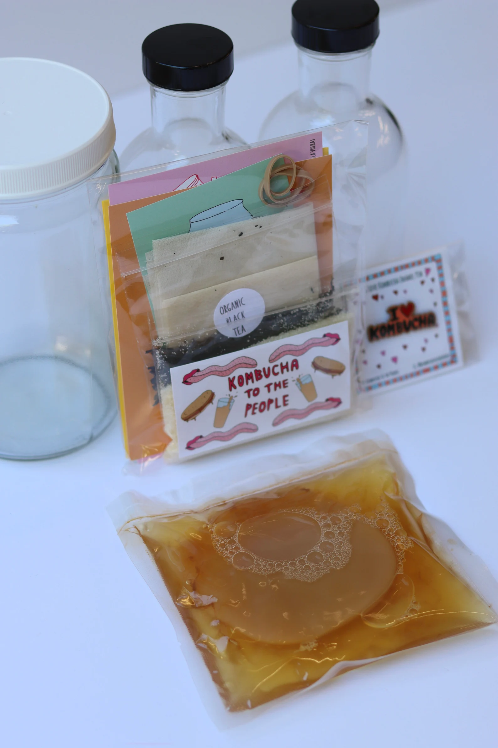 Kombucha brewing kit including glass jars, organic black tea, rubber bands, instruction cards, and a sealed pouch containing kombucha SCOBY and liquid.