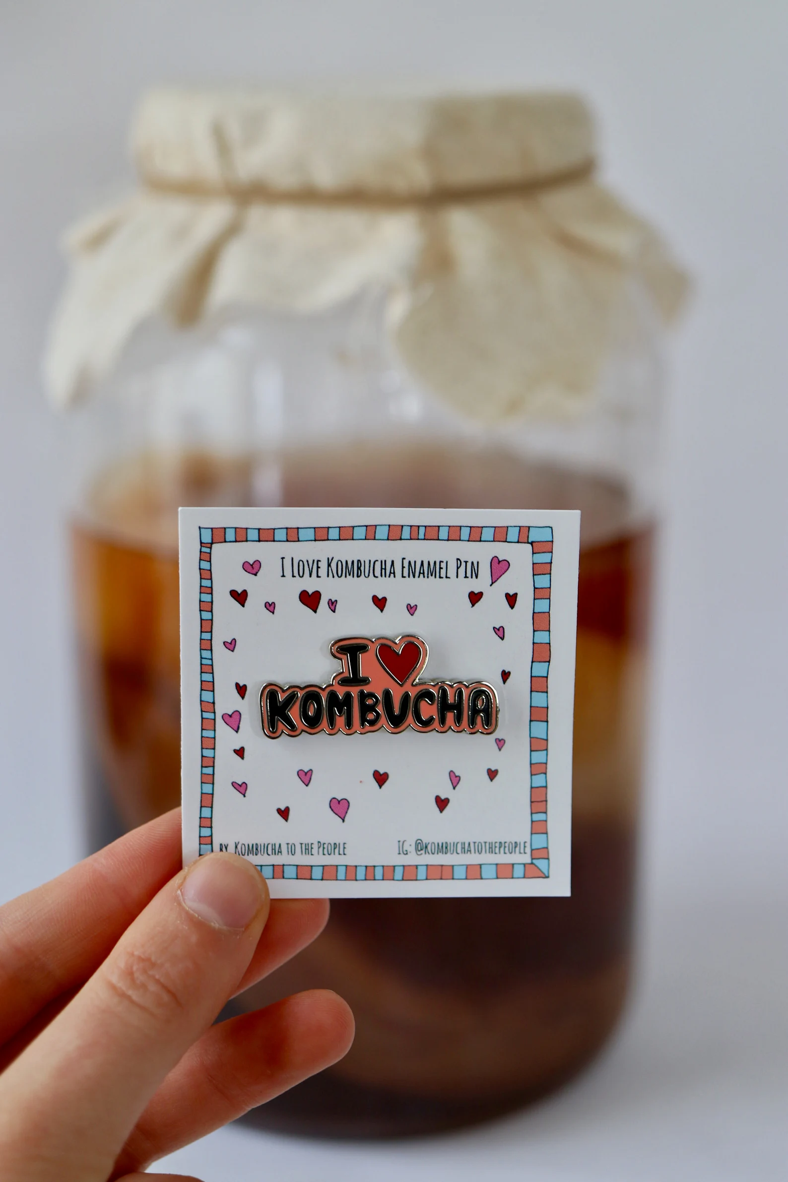 Hand holding an enamel pin on a card that says 'I ♥ KOMBUCHA' with hearts around it, with a jar covered by cloth in the background.