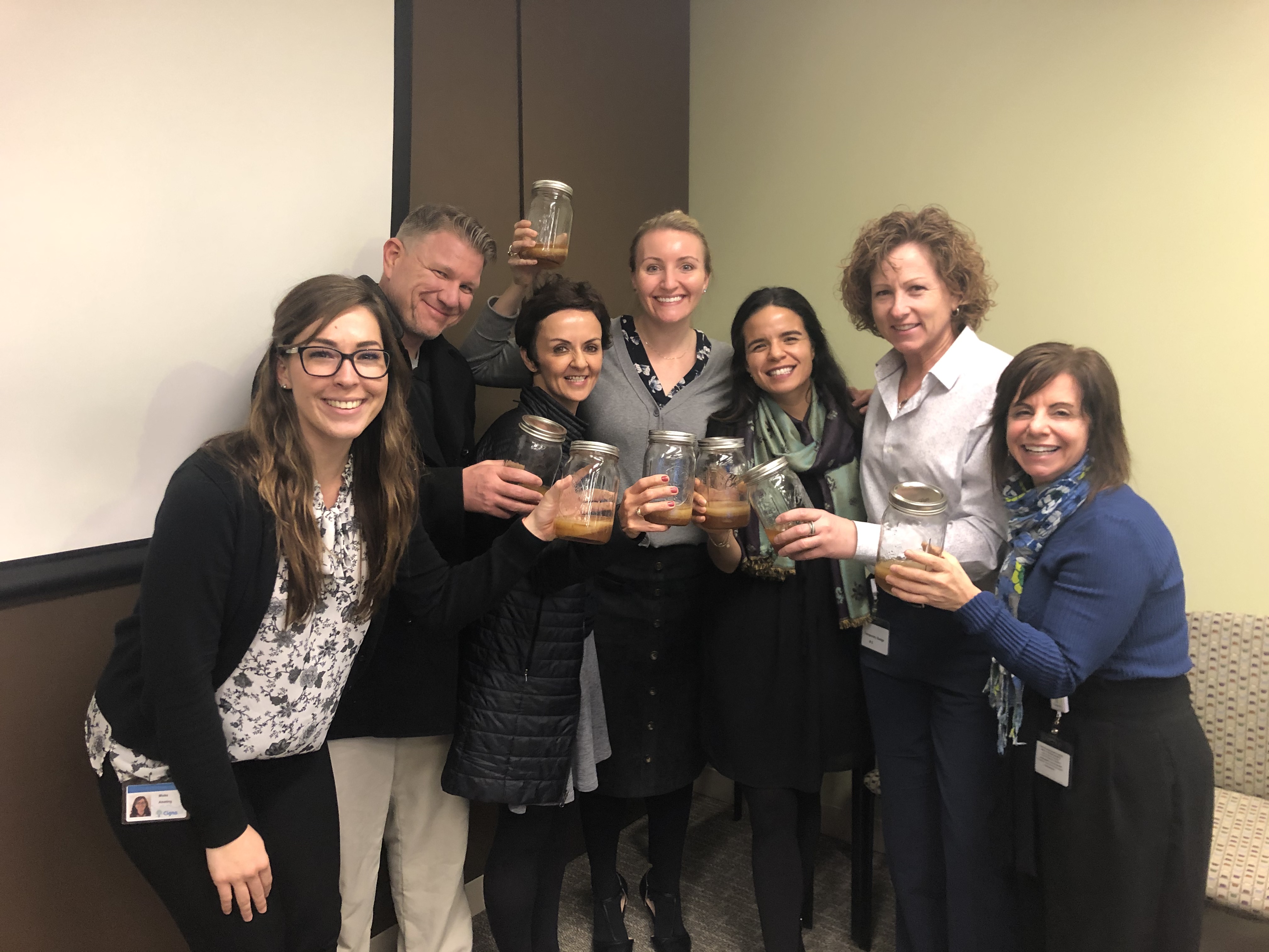 A group of seven smiling people holding up jars with a beverage, standing indoors against a beige and brown wall.