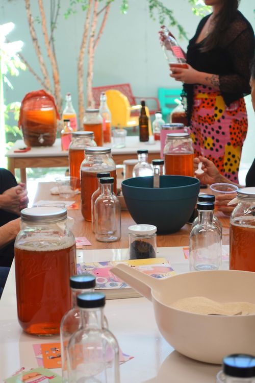 Table set with jars of kombucha, bottles, bowls, and ingredients, with people engaged in a kombucha brewing workshop.