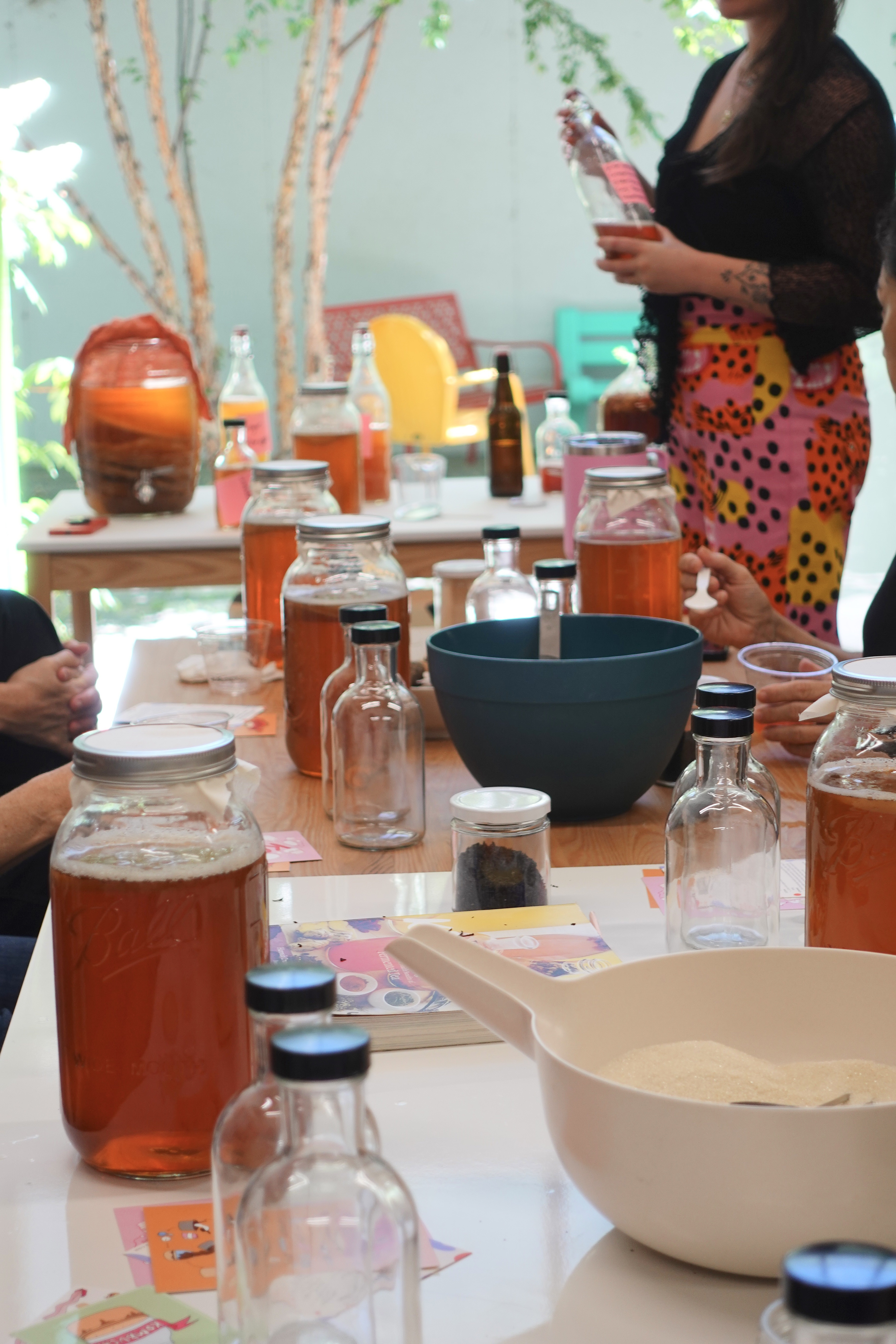 Table set with jars of kombucha, bottles, bowls, and ingredients, with people engaged in a kombucha brewing workshop.