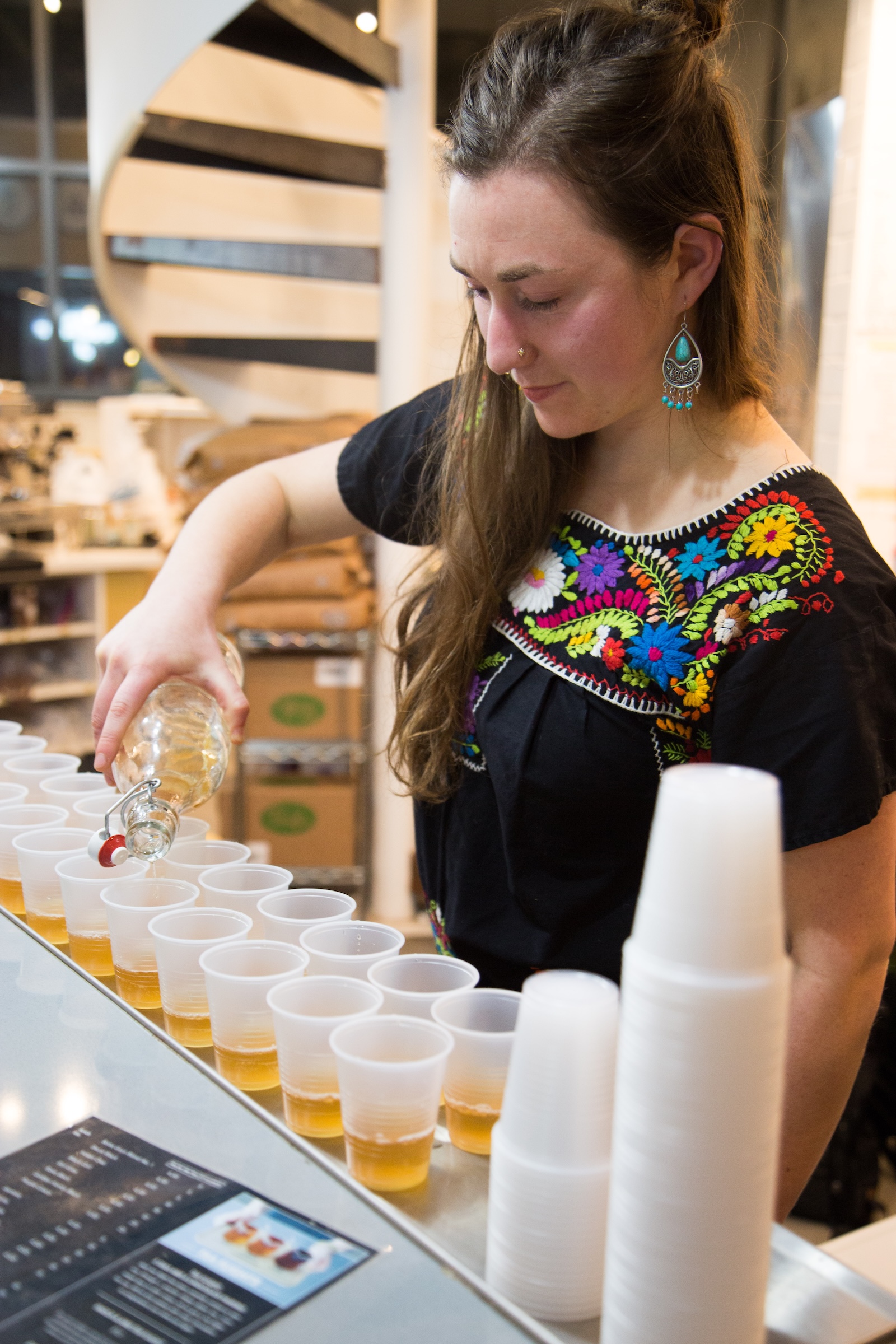 Woman in a black floral embroidered top pouring a kombucha beverage into a row of plastic cups on a counter.