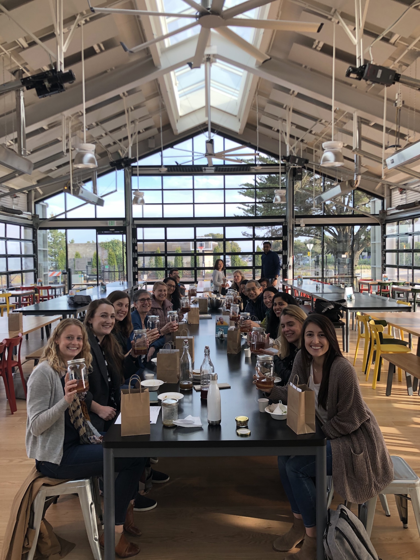 Group workshop participants seated at a long table holding their kombucha jars and smiling in a bright industrial event space
