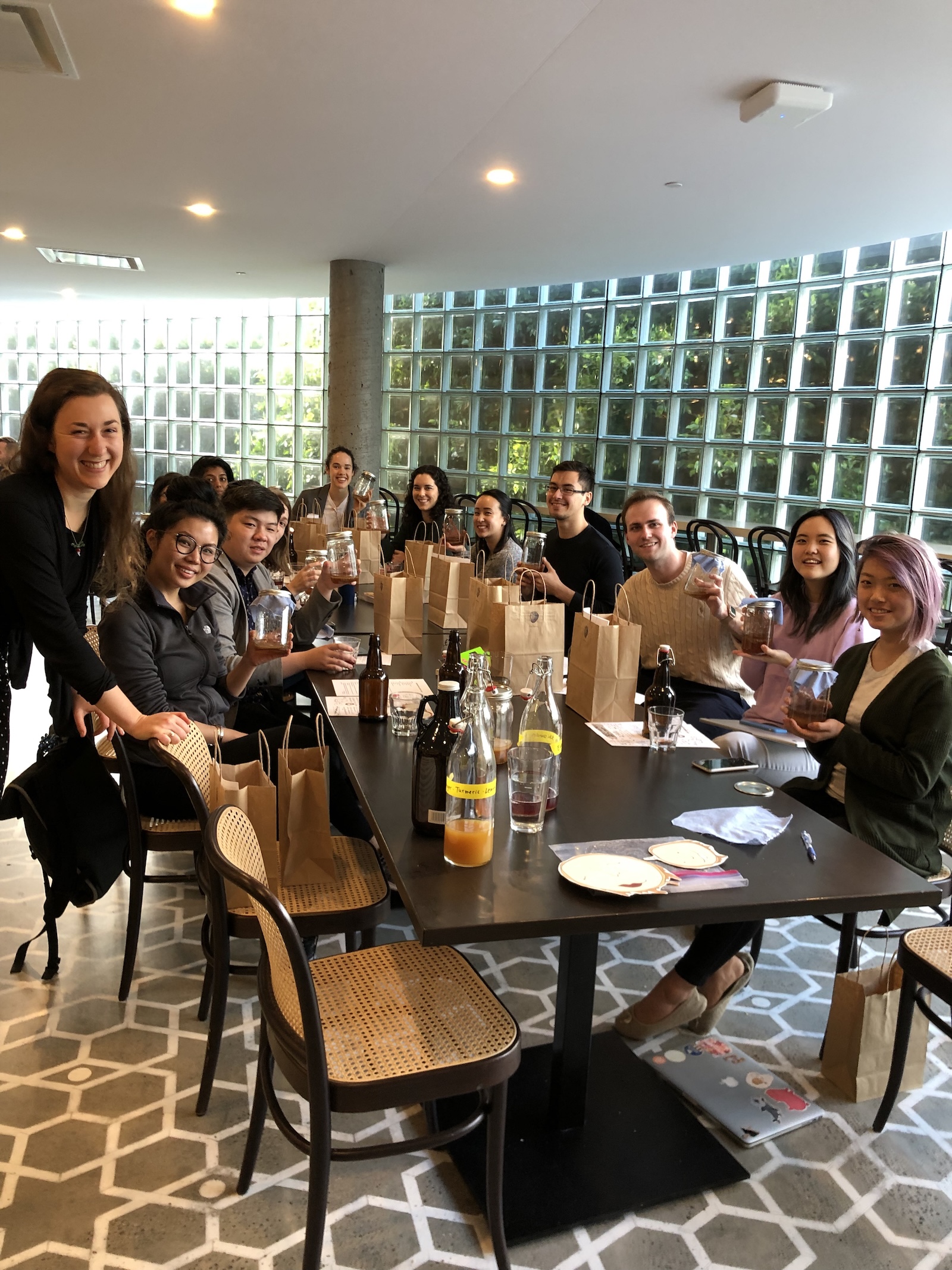 Group of young adults sitting around a long table holding kombucha jars, smiling during a social or workshop event with brown paper bags and bottles on the table.