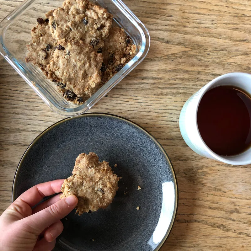 Homemade crust cookies stored in a glass Tupperware with a cup of tea on the side, as a hand holds a cookie piece, all set on a warm wooden countertop