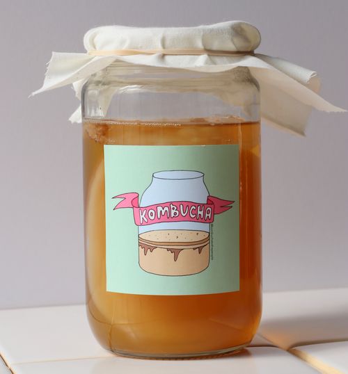 Woman presenting kombucha in a mason jar during a virtual corporate team building kombucha brewing class, set against the backdrop of a cozy kitchen.