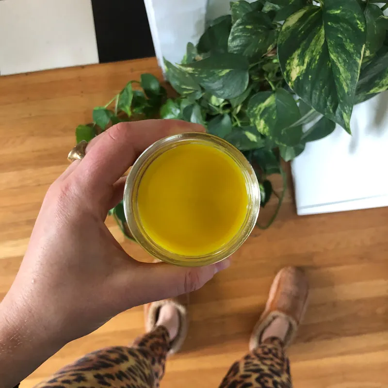 Hand holding a small glass cup with ginger and turmeric shot with a green plant behind and wood floor.