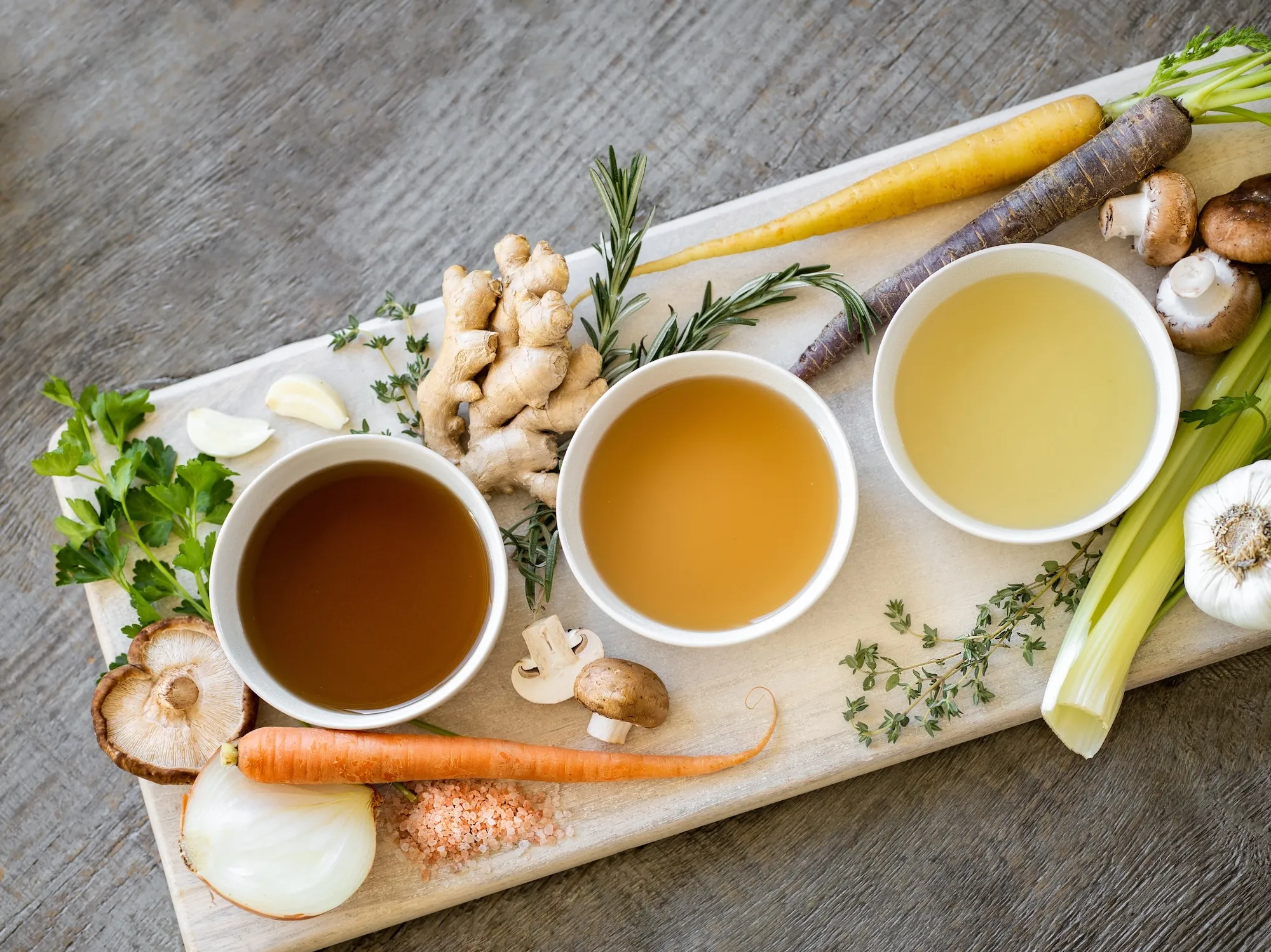 Three colorful bone broths in white bowls arranged on a wooden cutting board, surrounded by fresh carrots, ginger, and other vegetables and herbs.