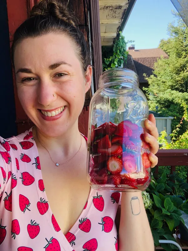 Woman wearing a charming strawberry print shirt, holds a mason jar filled with fresh strawberries.