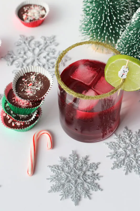 A red mocktail with a green rim, garnished with a lime wedge and peppermint chocolate, decorated with Christmas trees and snowflakes.