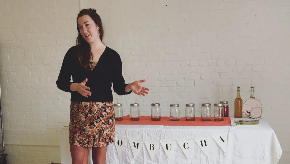 A woman teaching a kombucha brewing corporate class, with jars of kombucha on a table in the background, creating an engaging and educational wellness workshop atmosphere.