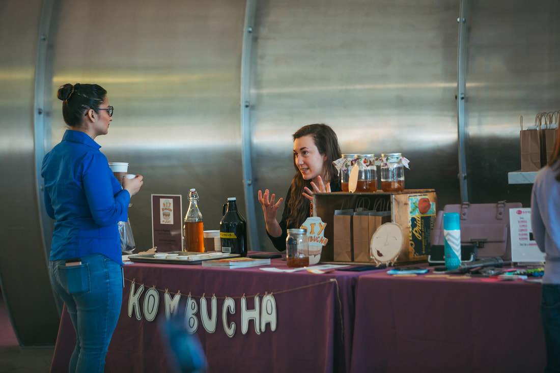 woman sitting at kombucha brewing table talking to another woman