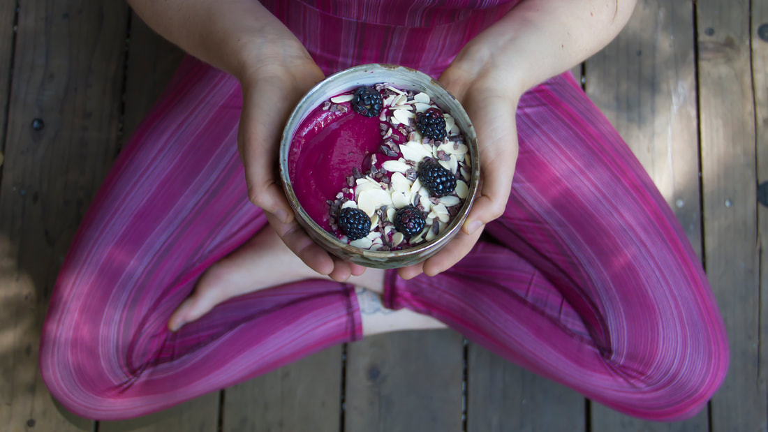 Kombucha smoothie bowl with vibrant berry blend, topped with blackberries, sliced almonds, and seeds, held in hands while seated outdoors on a wooden deck.