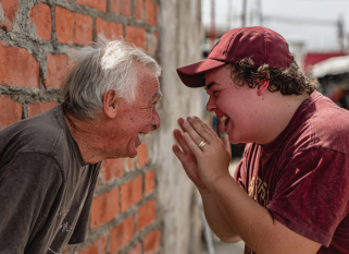An elderly man and a young man are laughing joyfully together while leaning toward each other near a brick wall. The young man, wearing a burgundy t-shirt and cap, clasps his hands in excitement, while the older man, dressed in a dark t-shirt, mirrors his smile. The scene conveys a genuine moment of shared happiness and connection.