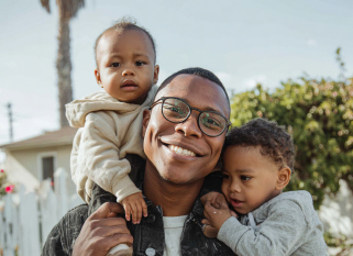 A smiling man wearing glasses and a black denim jacket holds two young children in his arms outdoors. One toddler sits on his shoulders wearing a beige hoodie, while the other child in a gray hoodie and red-striped pants clings to him lovingly. They are standing in front of a white picket fence with greenery and a house in the background on a sunny day.
