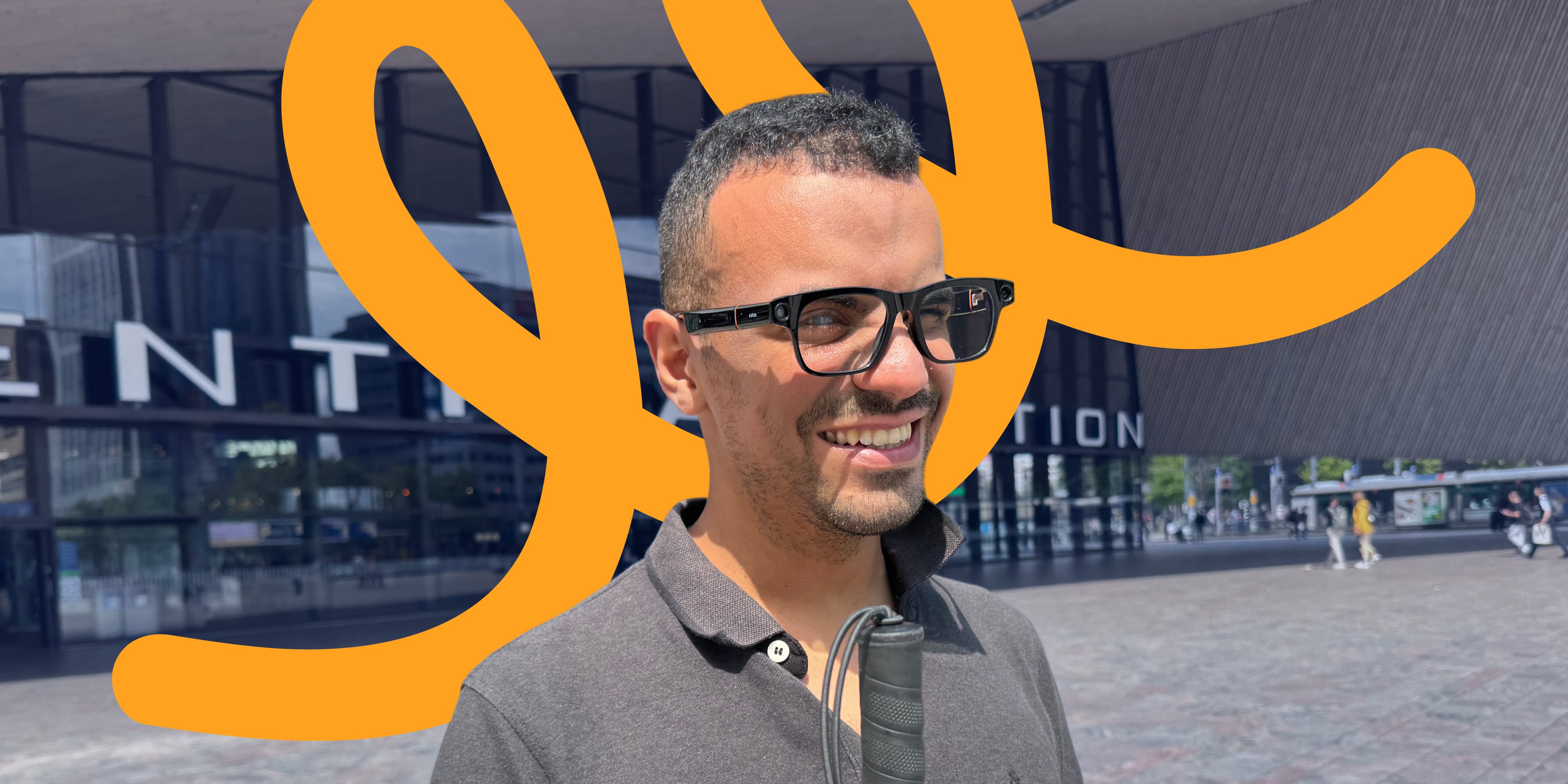 Smiling man wearing stylish black Solos smart glasses and a dark polo shirt, holding a white cane, standing outside a modern train station. An abstract orange swirl graphic overlays the background.