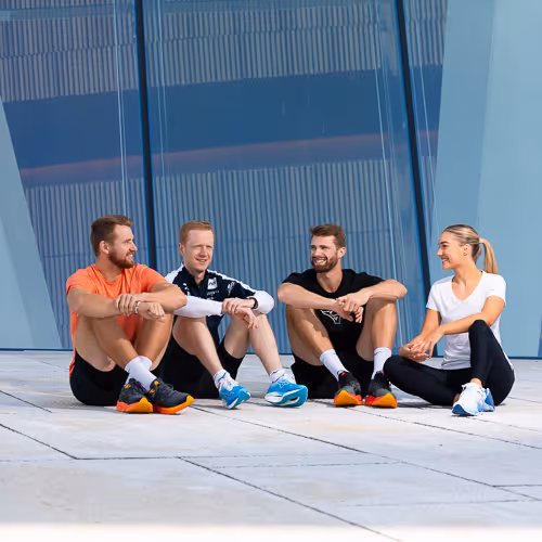Fire sporty kollegaer i treningsklær slapper av foran moderne glassfasade etter økt – sportsfotografering og videoproduksjon Oslo.