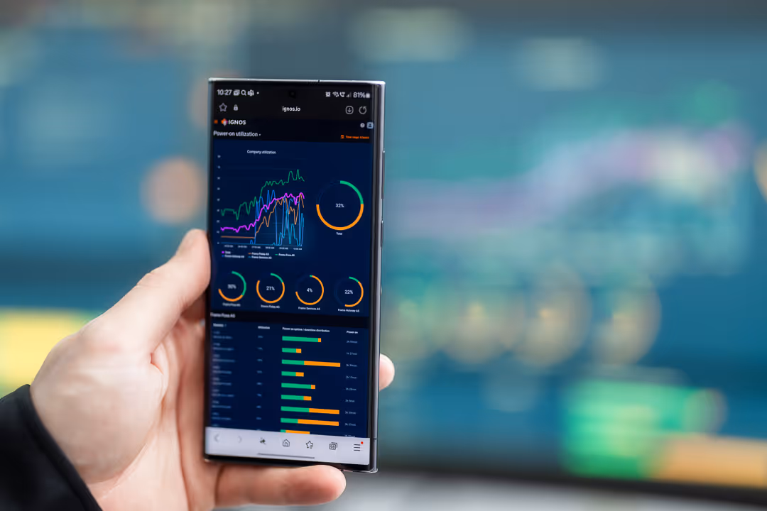 Hånd holder smart­telefon med produksjons­data-app foran uklar maskinlinje – industrifoto Stavanger.