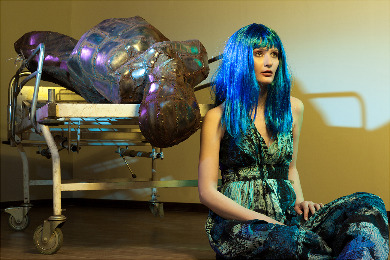 Conceptual fashion editorial of a blue-haired model seated among metallic and glass props
