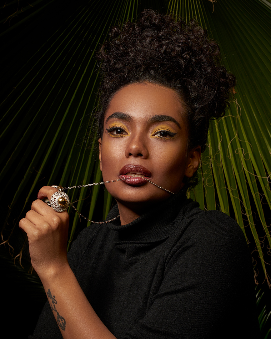 Editorial portrait of a model with an updo holding a ring near her face against dark green palm leaves