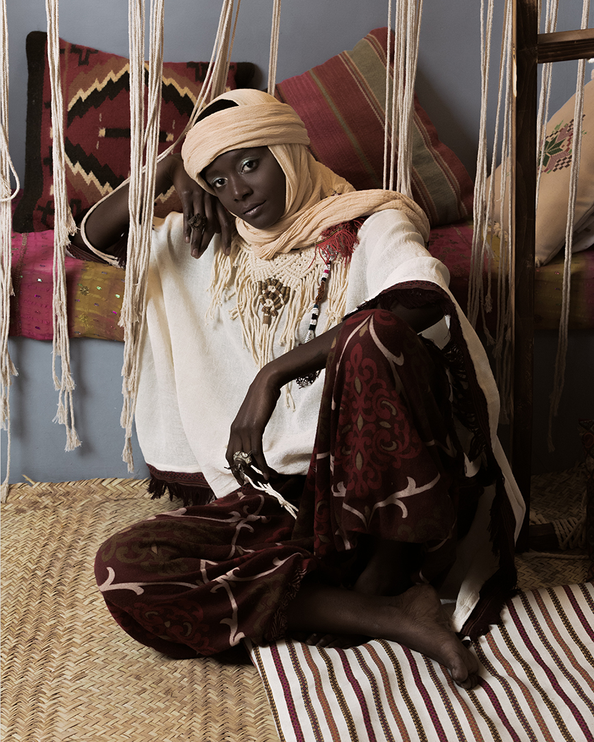 Editorial portrait of a model in traditional cream robes and a head wrap seated against a wall