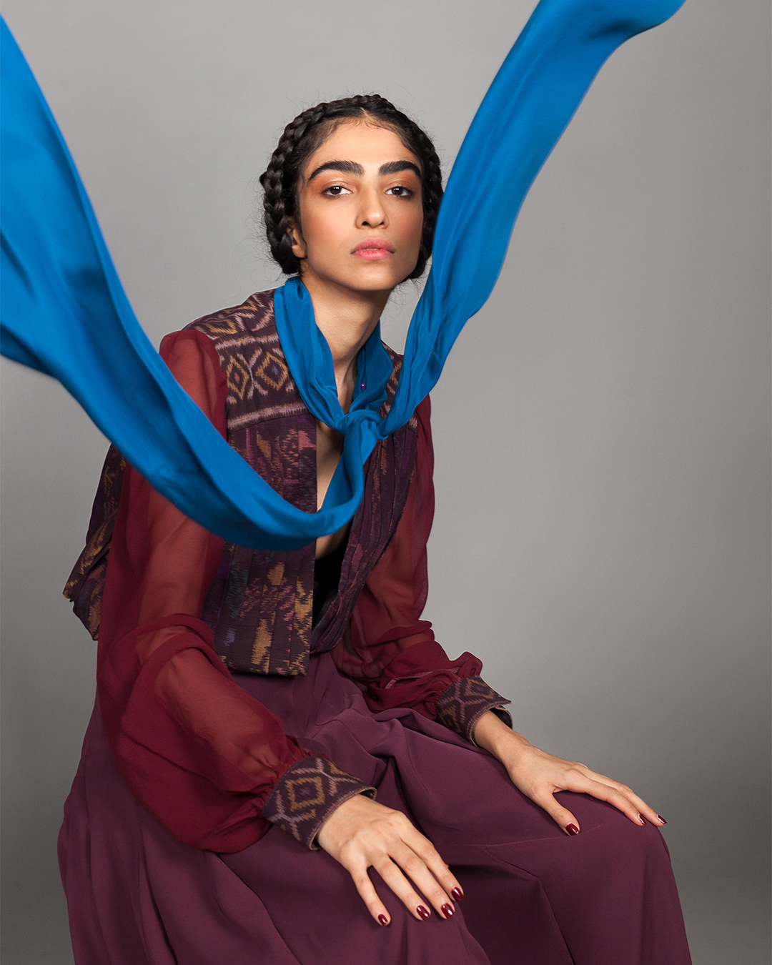Fashion portrait of a seated model in a burgundy dress draped with a blue silk scarf
