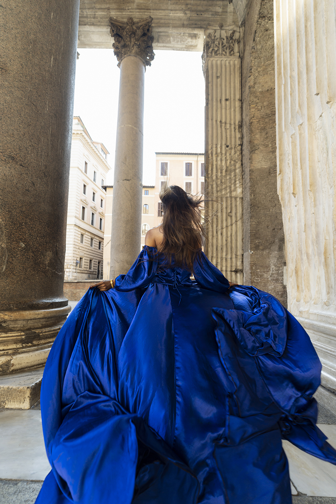 Royal-blue flying dress at the Pantheon colonnade, back view with wind-tousled hair and travertine columns, Rome.