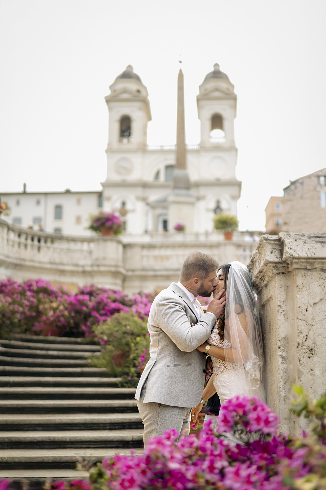 Bride and groom kissing among pink flowers at the Spanish Steps Rome, captured by Eidos Photography Rome photographer Jeff during a romantic morning wedding photoshoot.