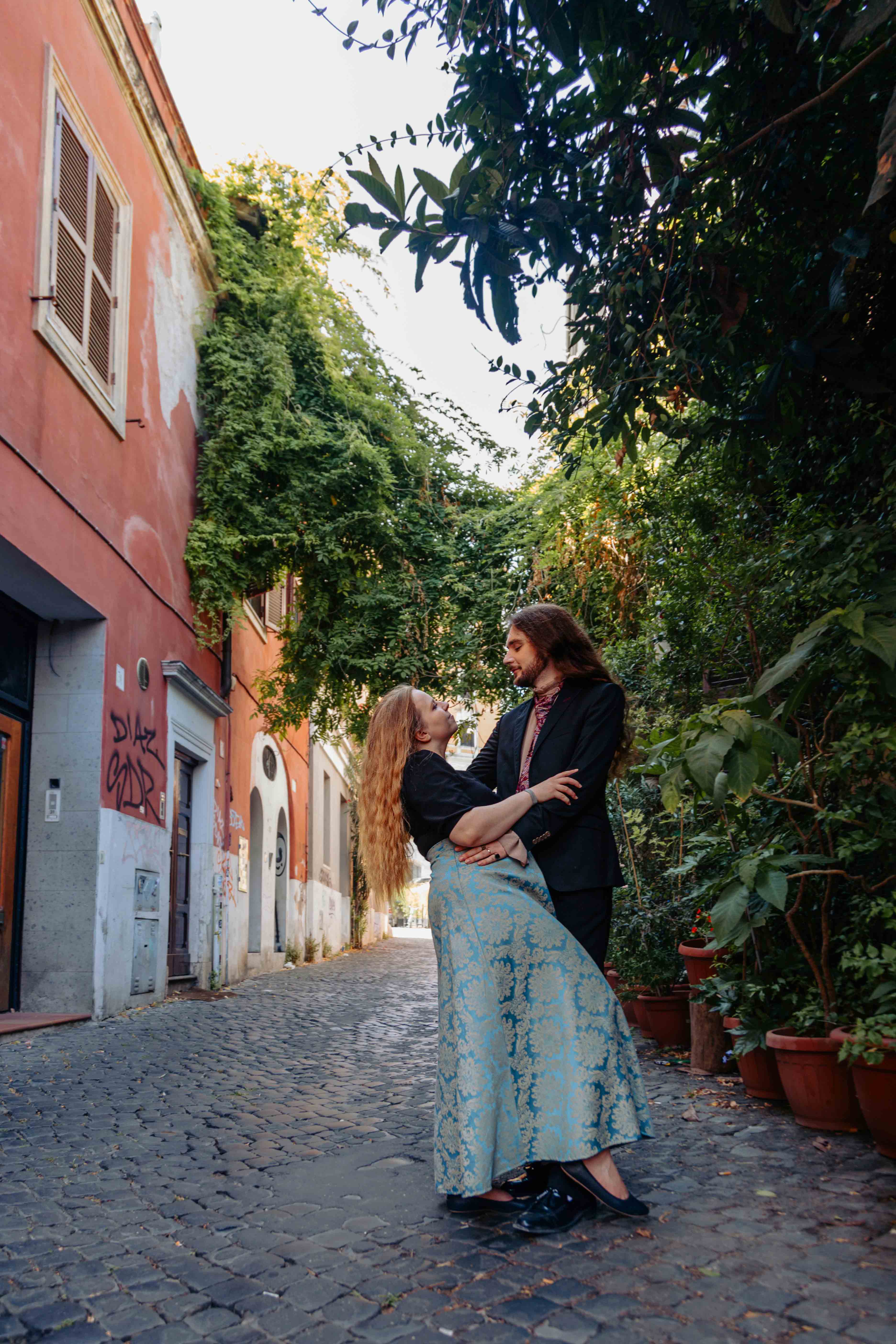 Romantic couple photoshoot in Trastevere, Rome, featuring two lovers embracing on a cobblestone street surrounded by colorful Italian buildings, captured by Eidos Photography.
