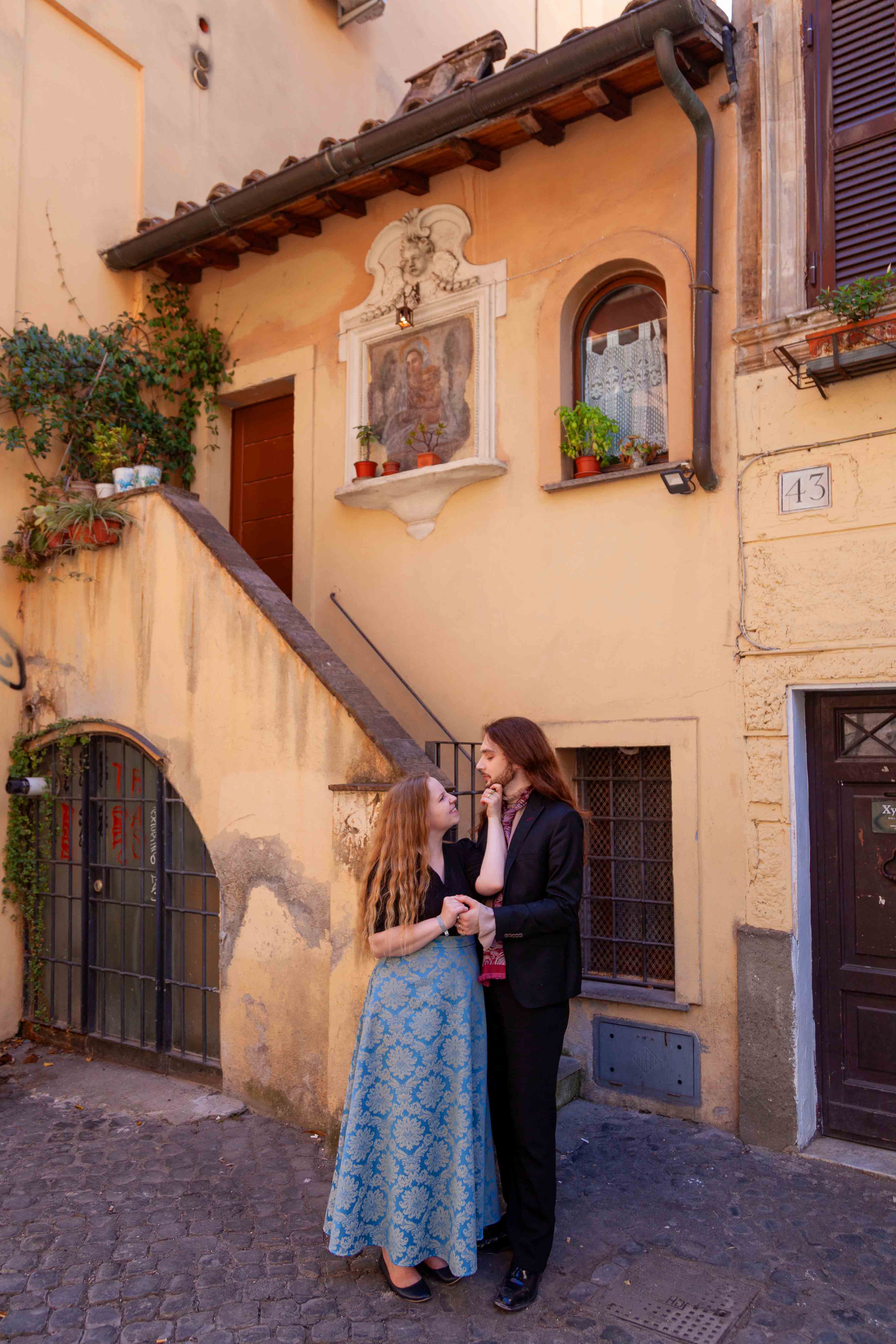 Couple photoshoot in Trastevere, Rome, with two lovers standing close on a cobblestone street beside colorful pastel buildings, captured by Eidos Photography.