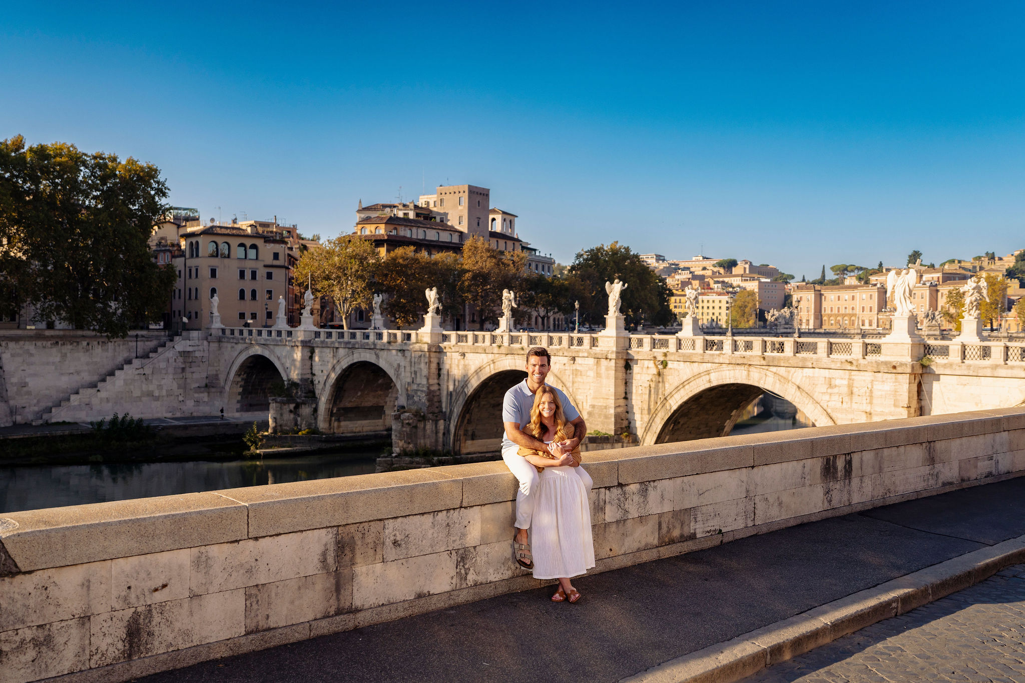A romantic couple sitting by the Tiber River in Rome with Ponte Sant’Angelo Bridge and classic Roman architecture in the background. Captured by Eidos Photography, this Vatican photoshoot spot offers golden evening light and a peaceful view connecting Vatican City and the heart of Rome.