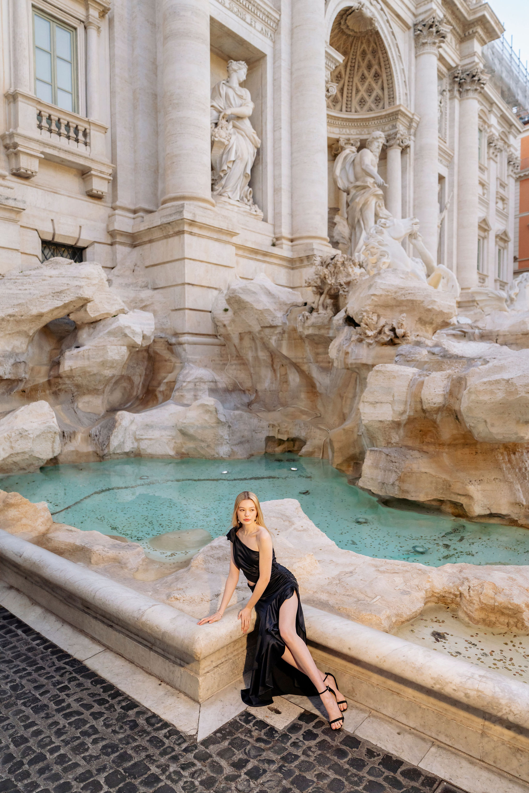 Solo portrait seated on the travertine edge by the Trevi Fountain pool before sunrise, clean cobblestones and soft blue-hour color, photographed by Eidos Photography Rome