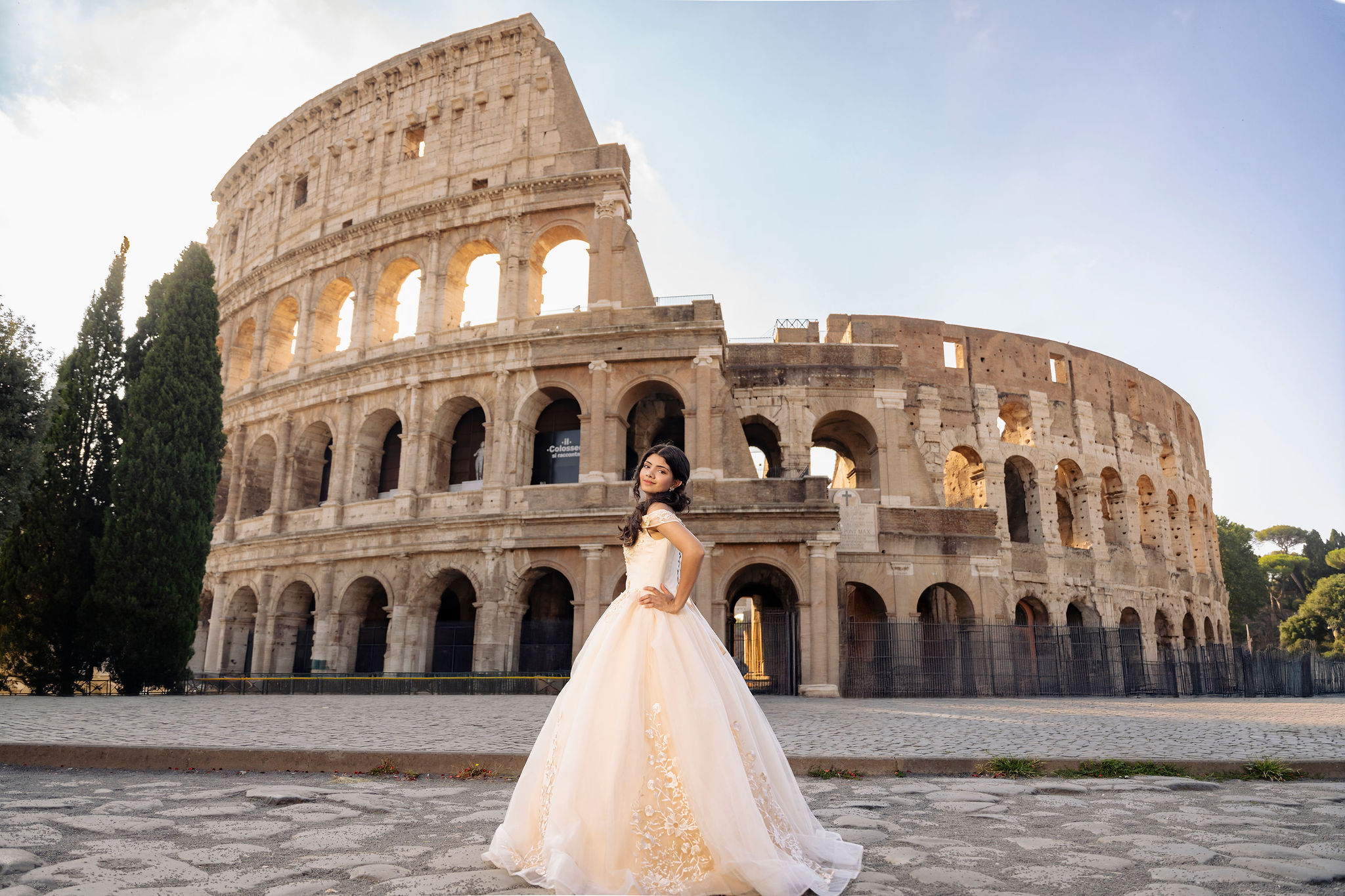 quinceañera in an ivory princess ball gown standing on a stone courtyard with the Colosseum in the background at first light in Rome