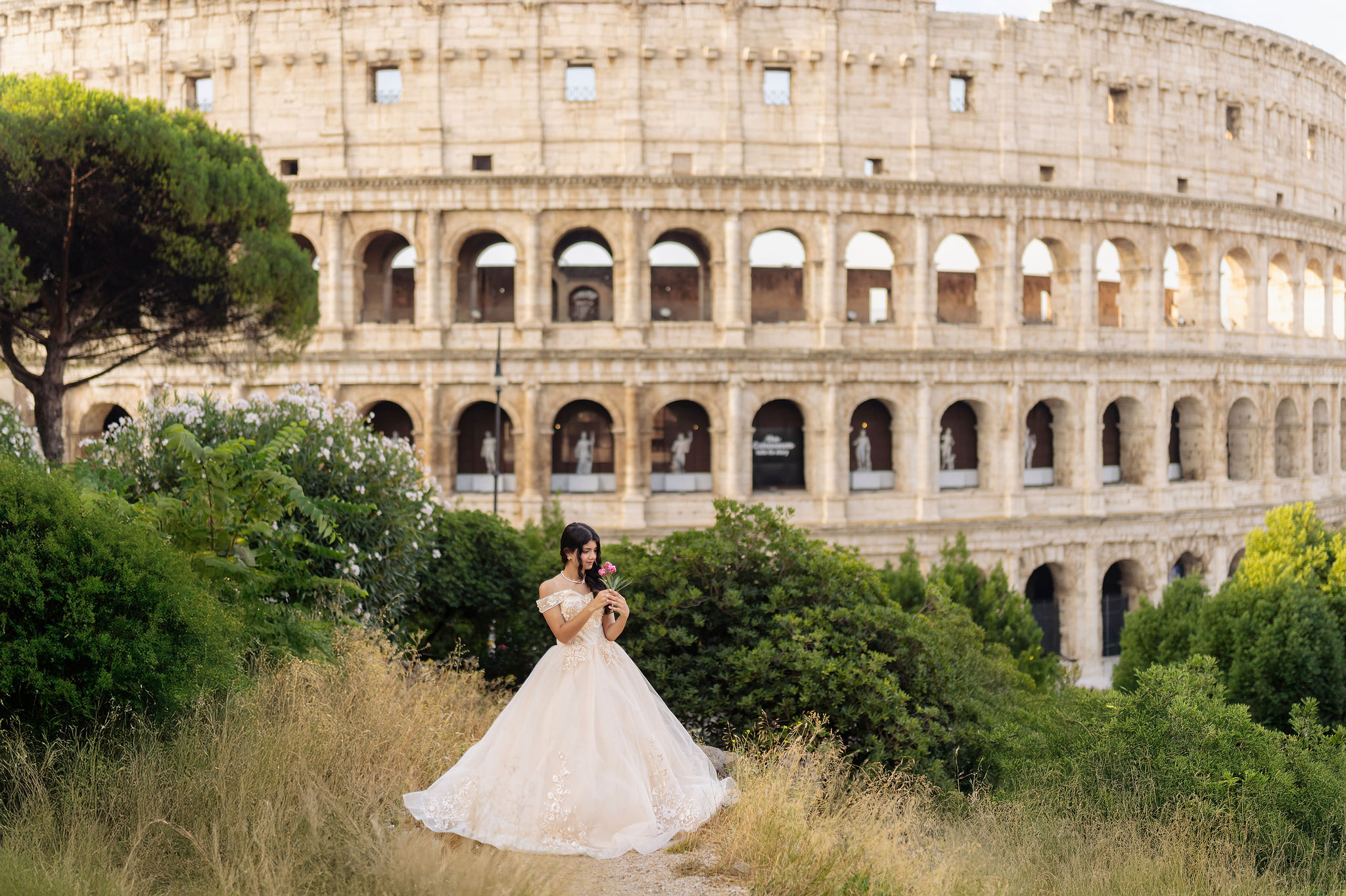 wide shot of a quinceañera in an ivory ball gown holding her dress on a grassy overlook with the Colosseum in the background at first light in Rome