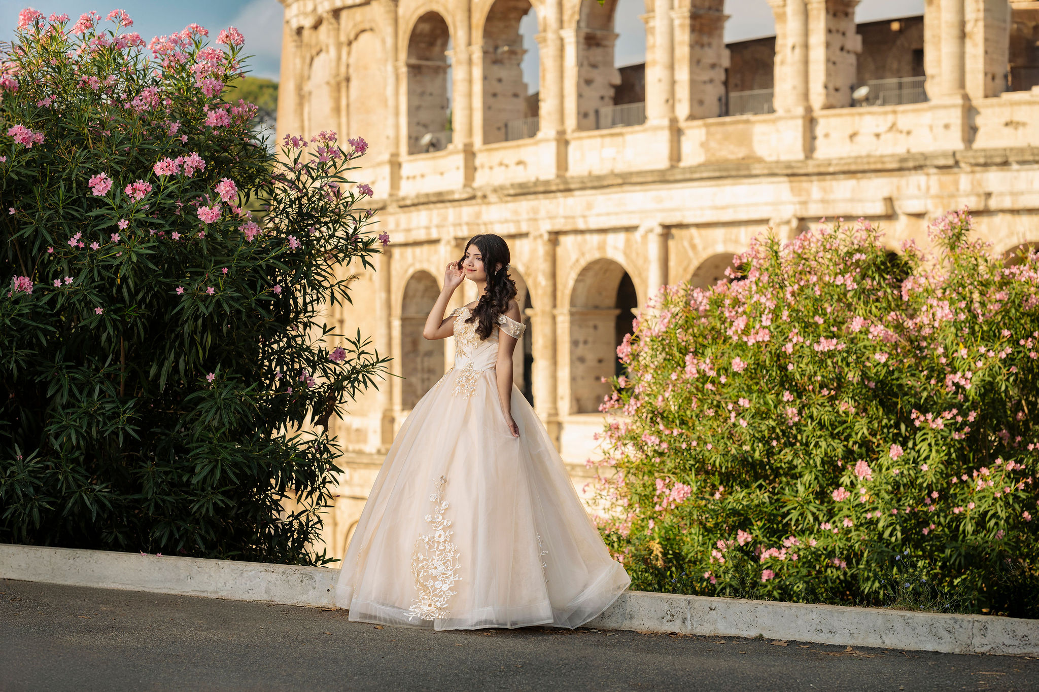 quinceañera in an ivory princess ball gown standing between pink flowering bushes with the Colosseum arches in the background at first light in Rome