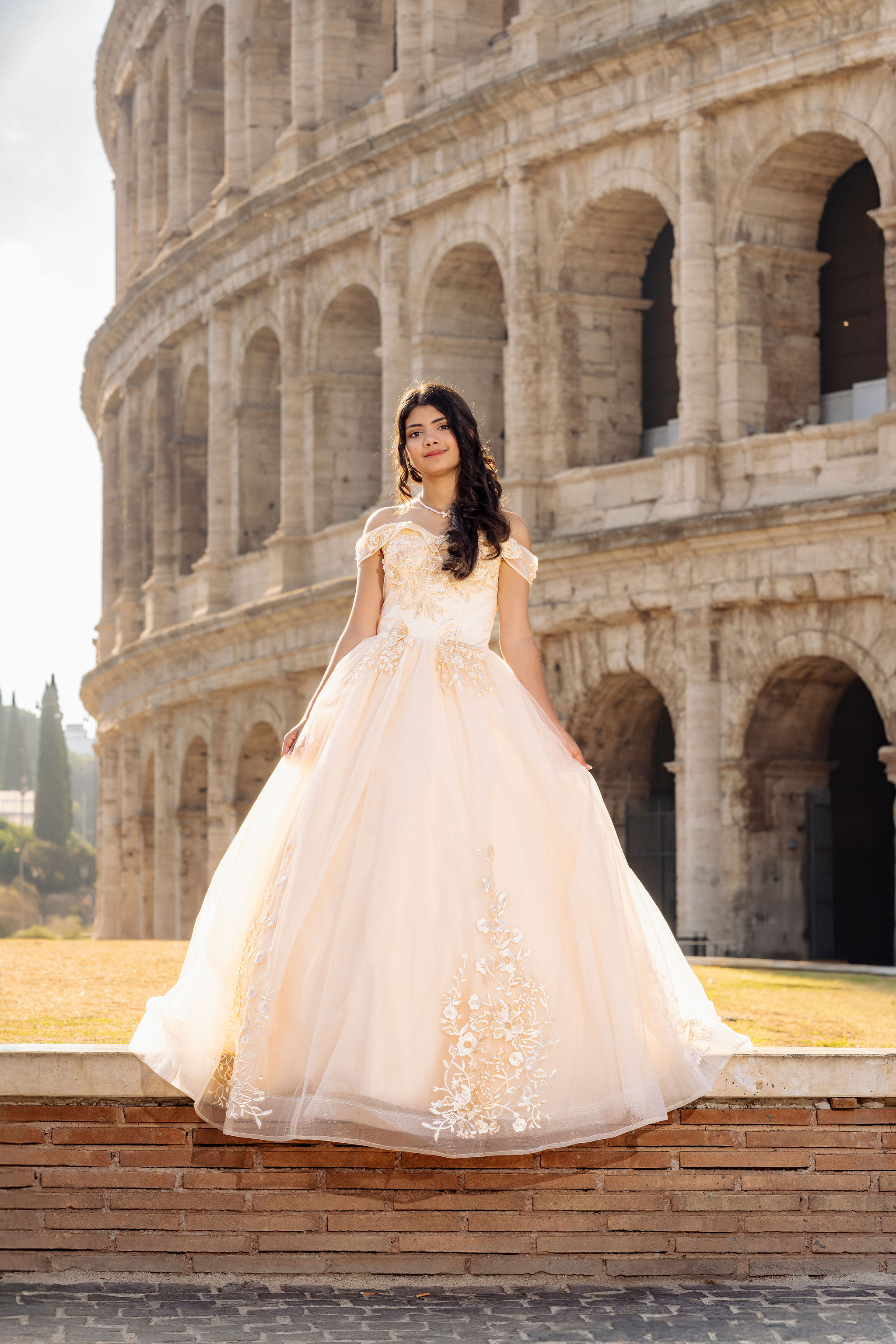 medium full portrait of a quinceañera in an ivory off-shoulder ball gown with lace details standing in front of the Colosseum at first light in Rome